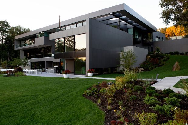 Exterior view of a large multi-level contemporary residence set within a landscaped suburban garden. The building features dark grey geometric volumes, extensive glazing, projecting balconies, a flat roof with a deep overhang, and a partially covered outdoor terrace with white furniture. A paved walkway crosses the lawn and planted garden toward the house.