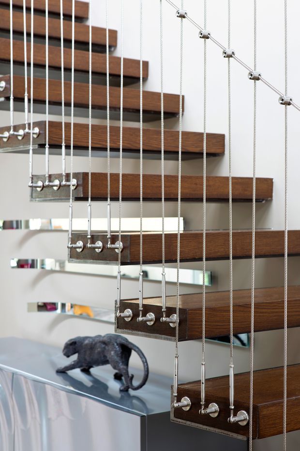 Close-up interior view of a sculptural floating staircase with dark timber treads suspended by numerous vertical stainless-steel cables and polished metal fittings. A reflective glass or metal console surface and decorative sculpture are visible below, with a clean light-coloured wall behind. The image emphasises the staircase construction and contemporary detailing.