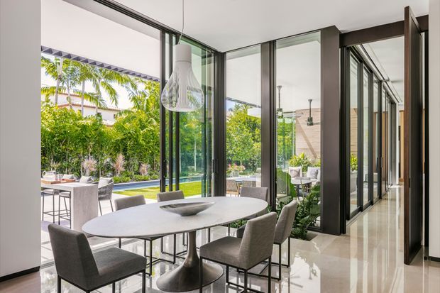 Bright contemporary dining area with a large oval table and upholstered chairs, opening through extensive black-framed glazing to a landscaped outdoor area and swimming pool. The space has polished light stone flooring, white walls, dark window and door frames, a sculptural pendant light, and a corridor of glazed openings extending to the right.