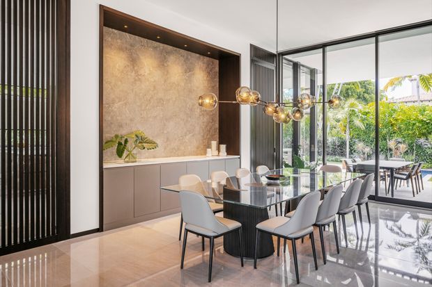 A refined contemporary dining room with a large rectangular glass dining table surrounded by upholstered dining chairs. A built-in wall display and storage unit features a warm textured stone-look backing, dark timber framing, integrated lighting, and lower cabinetry. Vertical dark slatted screens, polished light flooring, sculptural globe pendant lighting, and expansive floor-to-ceiling glazing create a bright connection to the landscaped outdoor area.