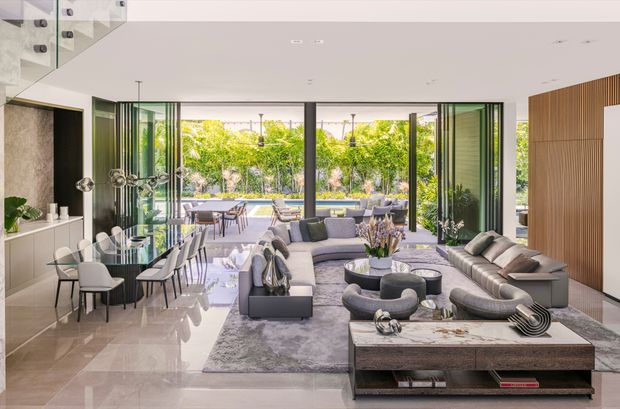 Open-plan contemporary living and dining area with a sophisticated neutral fit-out. The room features floor-to-ceiling glazing opening to an outdoor terrace and pool, a large sectional sofa arrangement, a dining table with sculptural pendant lights, polished light stone flooring, a marble-topped coffee table, dark built-in cabinetry, and a vertical timber feature wall.