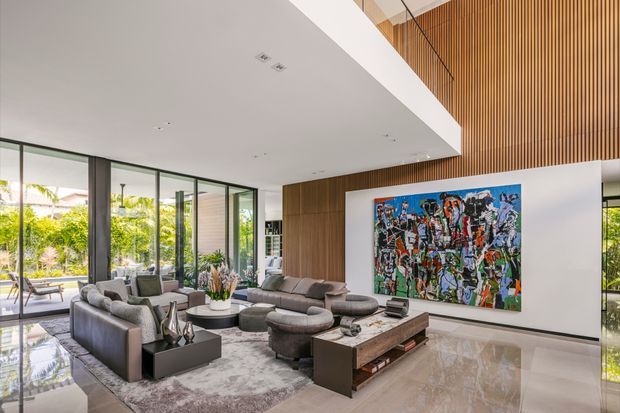 A spacious contemporary living room with double-height volume, extensive floor-to-ceiling glazing, and views onto landscaped greenery. The room features a large grey sectional seating arrangement, a low dark coffee table, a patterned area rug, polished pale stone flooring, a timber-panelled feature wall, and a vertically slatted timber ceiling or upper wall. A large colourful abstract artwork provides a strong focal point.