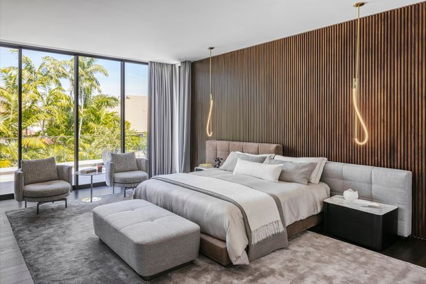 A sophisticated contemporary bedroom with a large upholstered bed, matching grey ottoman, two lounge chairs, floor-to-ceiling corner glazing, full-height curtains, and a dramatic dark timber slatted feature wall. Sculptural pendant lights hang beside the bed, while tropical greenery is visible outside.