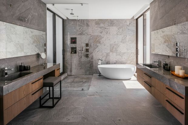 A spacious contemporary bathroom with a freestanding white oval bathtub, dual timber-veneer vanity units, large mirrored wall panels, and extensive grey stone-look wall and floor surfaces. Full-height glazing and a frameless glass shower enclosure bring in natural light. The room has a refined, minimalist-luxury feel with dark metal accents and linear ceiling spotlights.