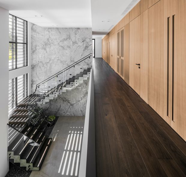 A bright, double-height interior stair hall with a long upper-level corridor. A dramatic staircase with dark treads and glass balustrades descends beside a large grey marble-look feature wall, while a full-height timber-lined wall conceals multiple doors and storage panels. Tall glazed openings bring in strong linear sunlight, highlighting the dark timber flooring and contemporary detailing.