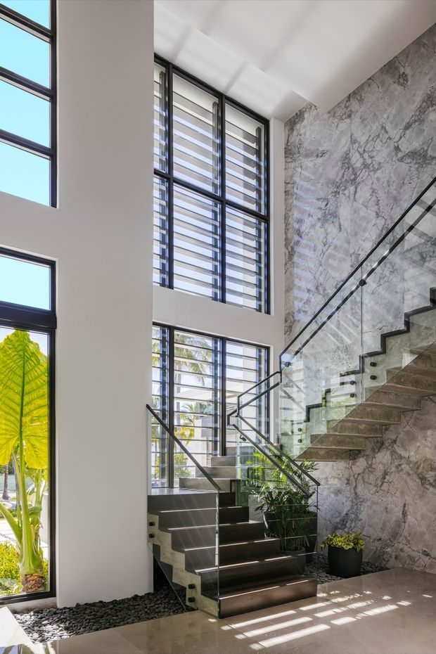 A bright, double-height interior stairway with dark timber or stone treads, clear glass balustrades, and slim dark metal handrails. Large black-framed louvred windows bring in abundant daylight and tropical greenery. One wall is clad in dramatic grey veined marble or stone, contrasting with smooth white walls and a pale polished floor. Decorative black pebbles and potted plants sit at the stair base.