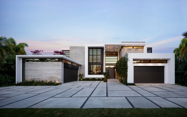 A contemporary multi-level residential facade viewed from the driveway at dusk. The home features crisp white rendered volumes, textured grey stone or concrete wall surfaces, extensive dark-framed glazing with horizontal louvres, warm recessed lighting, a prominent garage, and a large grid-patterned concrete driveway. Tropical landscaping frames the property.