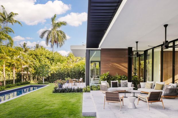 A bright contemporary outdoor entertaining area beside a modern home, with a covered patio furnished with lounge chairs, sofas, and small tables. The patio opens onto a manicured lawn and landscaped tropical garden, with a narrow rectangular swimming pool visible to the left. The building features broad glazing, dark slender structural posts, warm timber wall cladding, a white soffit, and a dark louvred roof-edge detail.