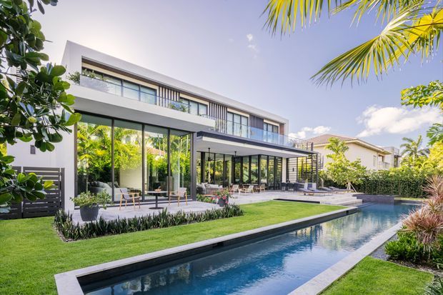 A contemporary two-storey residence with clean white rendered volumes, extensive floor-to-ceiling glazing, dark framed windows, and a generous covered outdoor living area. The home opens onto a manicured lawn and a long narrow swimming pool, with tropical planting and palm trees creating a resort-like setting.
