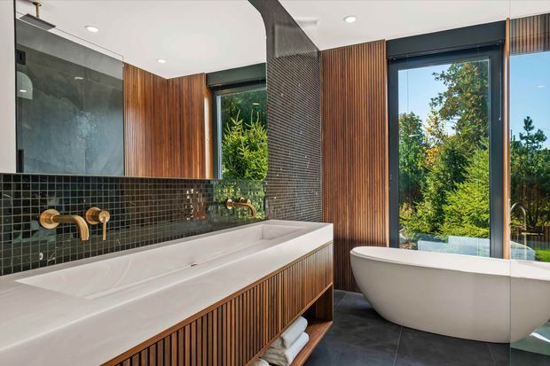 Contemporary bathroom featuring a long white double vanity with ribbed timber cabinetry, dark green square-tile splashback, wall-mounted brass fittings, a freestanding white bathtub, dark floor tiles, and extensive glazing overlooking greenery. Vertical timber wall panelling and a textured dark feature wall add warmth and contrast.