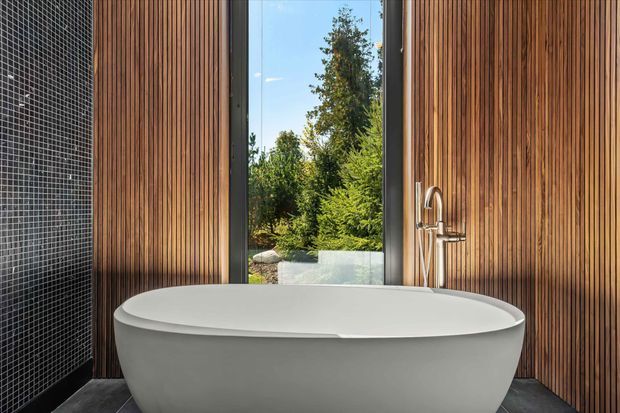 A contemporary bathroom featuring a freestanding white oval bathtub positioned against a richly toned vertical timber-slatted wall. A large narrow window opens to a leafy garden view, while a chrome floor-mounted bath filler stands beside the tub. The left wall is clad in small dark mosaic tiles, and dark flooring reinforces the high-contrast, refined palette.