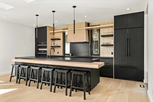 A spacious contemporary kitchen with a large island and seating for five or more. The room features light timber flooring and cabinetry, matte black full-height joinery, black benchtops, open shelving, integrated appliances, and slim black pendant lights. Bright white walls and ceiling create a clean, high-contrast interior.