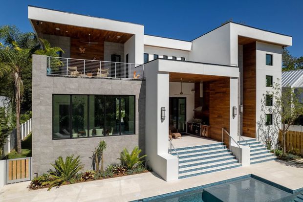 Contemporary multi-level white-rendered home with strong geometric volumes, flat rooflines, recessed outdoor living areas, and extensive glazing. Warm timber-lined ceilings and walls contrast with pale stone cladding, while broad stepped entry stairs lead toward a swimming pool in the foreground. Tropical planting and palm trees frame the facade.