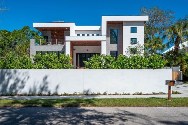 A contemporary multi-storey residence viewed from the street, with crisp white rendered volumes, flat rooflines, extensive dark-framed glazing, warm timber soffits and recessed wall sections, and a front balcony. Dense tropical landscaping sits behind a tall white boundary wall, with a driveway and letterbox visible at the right.