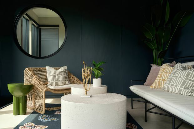 A moody, stylish living room with deep charcoal walls, a white upholstered sofa, woven rattan armchair, round pale terrazzo coffee table, and a small green side table. Patterned cushions, indoor plants, sculptural dried branches, and a large round wall mirror add texture and visual interest. The space has a compact, refined contemporary feel with strong contrast between dark walls and light furnishings.