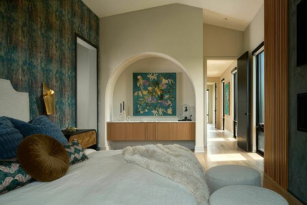 A sophisticated bedroom with a bed in the foreground, deep teal textured feature walls, and a pale neutral ceiling and floor palette. An arched recessed niche frames colourful artwork above a long timber-finish console, while a bright hallway extends beyond the room. Vertical timber battens and dark built-in elements line the right wall, with layered cushions and soft bedding adding comfort and richness.