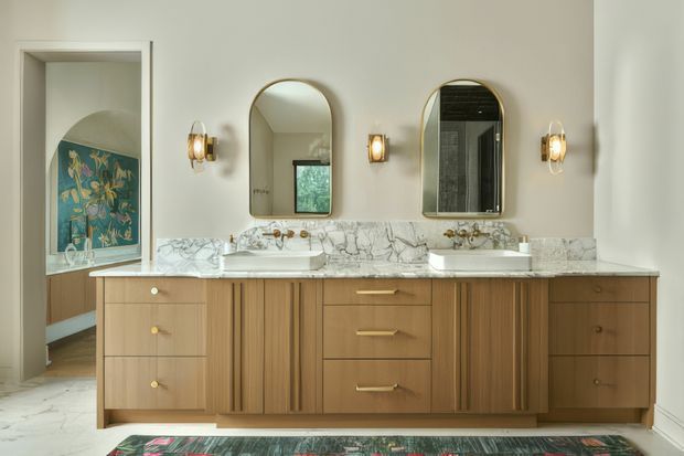 Elegant double-vanity bathroom with a long bespoke timber vanity, twin white basins, white veined marble benchtop and matching marble splashback. Two arched mirrors and warm brass wall sconces sit above the vanity. A doorway at left reveals part of an adjacent bedroom or sitting area, while patterned artwork and a rug add colour and character.