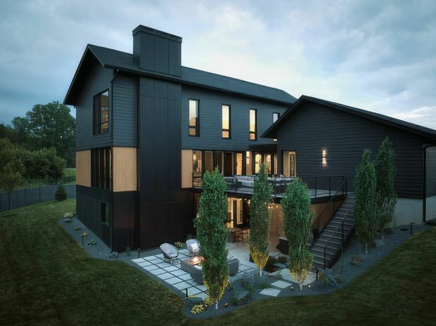 Exterior architectural view of a dark-clad multi-level house on a sloped suburban site. The home combines a tall gabled volume with a projecting lower side wing, dark horizontal and vertical cladding, a pitched roof, prominent chimney, tall narrow windows, and an elevated terrace with stairs descending to a paved courtyard. Landscaping and slender trees surround the outdoor living area.
