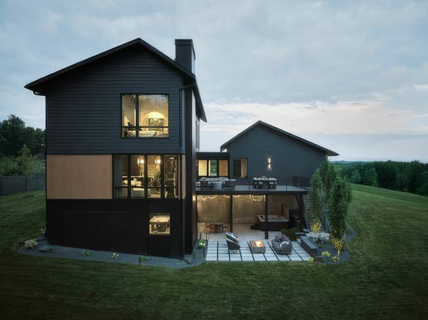 Exterior view of a contemporary dark-clad multi-level house on a spacious grassy site. The home has steep gable roofs, charcoal horizontal cladding, timber-toned facade panels, extensive glazing, and a partially sunken lower level opening onto a paved outdoor terrace with seating and a fire feature. Landscaping and distant rolling countryside surround the residence.