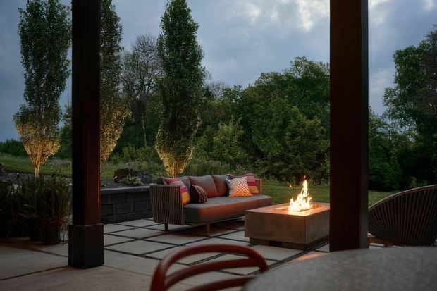 A covered outdoor terrace overlooking a landscaped garden, framed by substantial dark timber posts. The space features a contemporary outdoor sofa with colourful cushions, a square built-in fire pit, tiled or paved flooring, low retaining walls, and additional outdoor seating. The scene has a warm firelit atmosphere beneath an overcast sky.