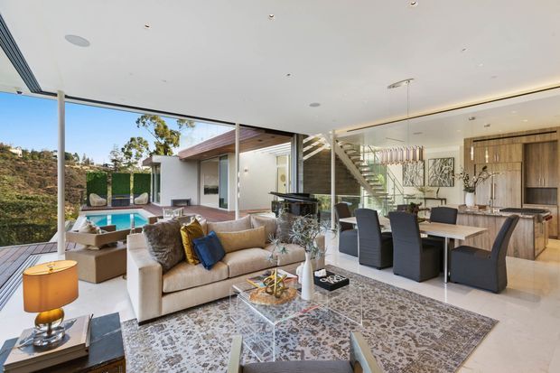 Open-plan contemporary living, dining and kitchen area with floor-to-ceiling glazing opening onto a pool terrace and landscaped hillside. The bright interior features a large sectional sofa, patterned area rug, glass coffee table, dining table with dark upholstered chairs, timber kitchen cabinetry, a sculptural pendant light, and a visible staircase leading to an upper level.