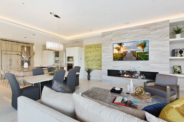 Bright open-plan contemporary living and dining area with a neutral palette. The room features a large sectional sofa, upholstered dining chairs around a timber dining table, a textured stone feature wall with a built-in black fireplace and wall-mounted television, recessed shelving, pendant lighting, and integrated kitchen cabinetry visible in the background.