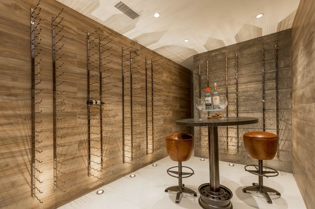 A compact, richly clad wine room with warm wood-look wall surfaces, extensive vertical wine bottle racks, and a small round bar table with two stools. The bright white floor includes recessed uplights, while the ceiling has patterned wood panels, recessed downlights, and ventilation grilles. A dark textured feature wall and illuminated bottle display add contrast.