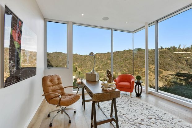 A bright, compact modern home office with floor-to-ceiling corner glazing overlooking a rugged wooded hillside. The room has white walls, pale timber flooring, a vintage-style leather swivel chair, a vivid orange-red lounge chair, a dark wood desk, a patterned light rug, artwork, books, and a small sculptural lamp. The space feels airy, refined, and connected to the landscape.