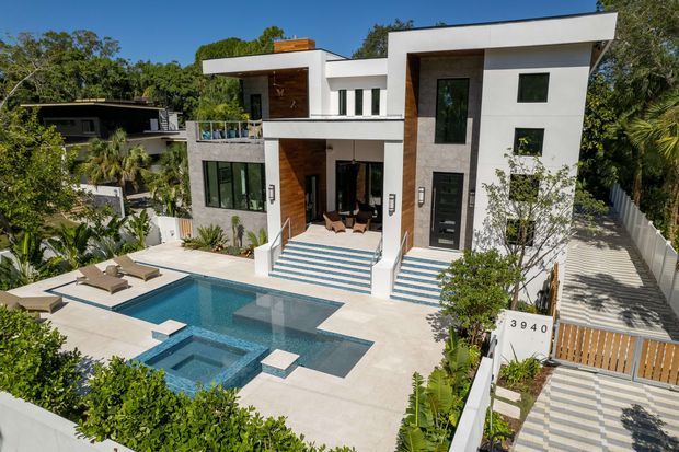 Exterior architectural photograph of a contemporary multi-level white residence with strong geometric forms, flat roofs, dark-framed windows, grey stone-clad sections, and warm timber wall accents. The home overlooks a tiled terrace with a swimming pool and spa, sun loungers, planted landscaping, and a patterned pedestrian entrance path.