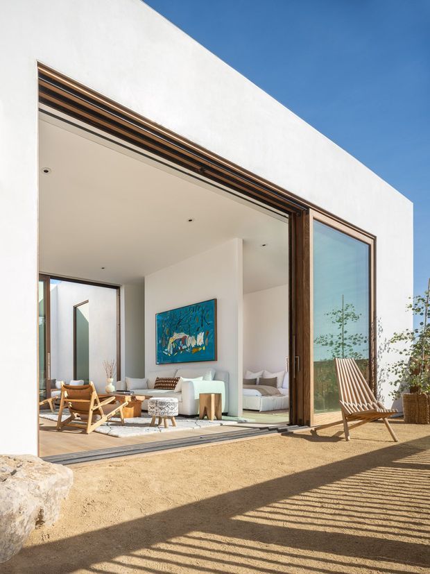 Bright architectural photograph of a minimalist white-rendered residence with large sliding glass doors opening directly onto a sandy outdoor area. Warm timber window and door frames, timber-lined overhead detailing, pale timber flooring, white furnishings, and a colourful wall artwork are visible inside. A timber slatted lounge chair sits outside, with strong sunlight and shadows creating a relaxed indoor–outdoor connection.