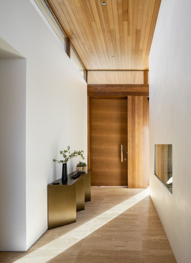 A minimalist residential entry hall with a tall timber-lined ceiling, warm wood pivot door and matching timber wall panel, pale stone or timber-look flooring, and crisp white walls. A slim dark-bronze console with restrained decor sits along the left wall, while a narrow internal window or opening appears on the right. Strong natural light creates a diagonal beam across the floor.