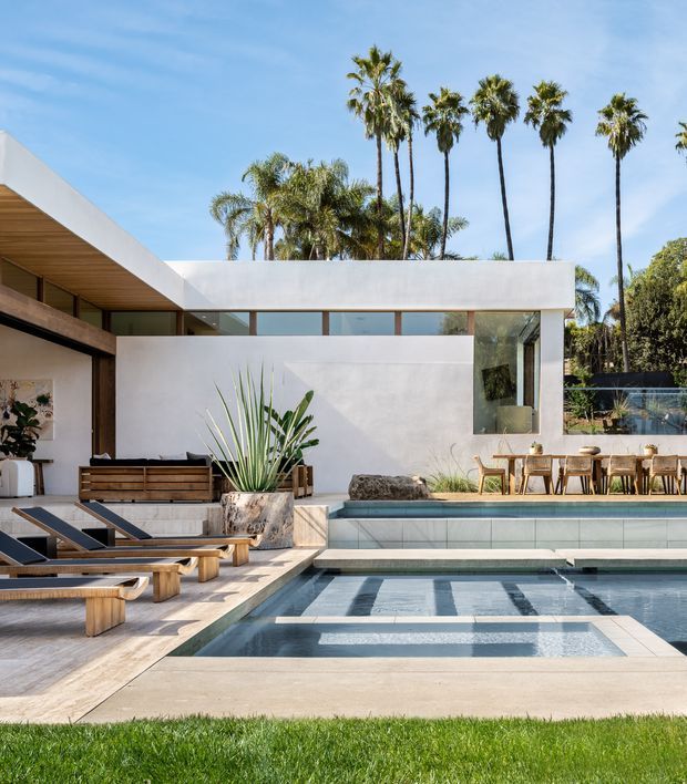 A modernist residential pool courtyard with a crisp white flat-roofed facade, extensive glazing, warm timber soffits and screening, and an adjacent outdoor dining area. The foreground features a rectangular swimming pool with integrated shallow steps, pale stone or concrete paving, timber sun loungers, and sculptural drought-tolerant planting. Tall palm trees rise behind the house under a clear blue sky.