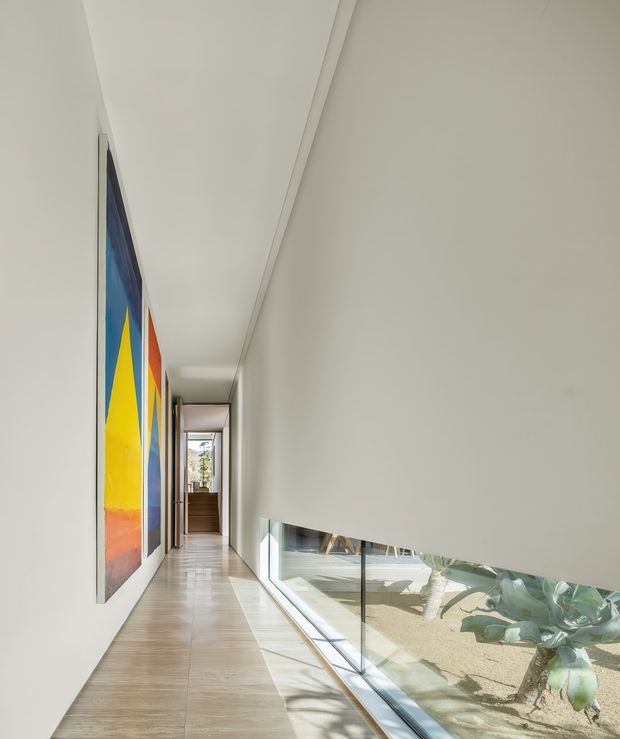 A long, minimalist interior corridor with pale tiled flooring, smooth white walls and ceiling, large colourful abstract artworks along the left wall, and a low horizontal glazed window on the right overlooking a planted exterior courtyard. Natural daylight creates a bright, calm atmosphere, with a doorway and additional glazing at the far end.
