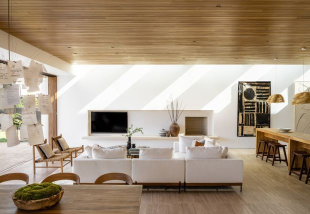 Bright open-plan living and dining area with a warm timber slatted ceiling, pale timber flooring, white built-in media joinery, large sectional seating, and a long timber dining table. Strong diagonal sunlight creates a dramatic pattern across the white wall, while sculptural artwork, natural decor, and woven lighting add warmth.