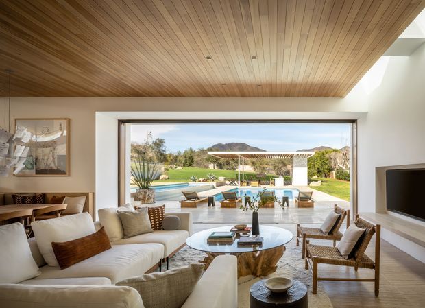A sophisticated open-plan living room with a warm timber-lined ceiling, pale walls, large picture-frame glazing, and a neutral seating arrangement around a round glass coffee table. The room opens directly to a landscaped backyard with a swimming pool, covered outdoor entertaining structure, and distant hills. Natural materials, woven chairs, soft upholstery, and abundant daylight create a relaxed contemporary indoor-outdoor setting.
