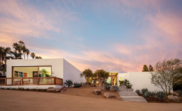 Low-rise modern white rendered building complex viewed from an open landscaped forecourt at sunset. The composition features crisp cubic forms, a large timber-framed window and timber balustrade on the left, a recessed central entrance reached by broad steps, and drought-tolerant desert planting with agaves, palms, sculptural trees, and rocks. The warm sunset sky creates a calm, resort-like atmosphere.