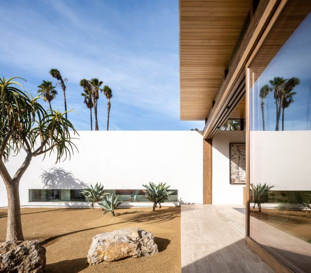 Exterior architectural view of a minimalist modern residence arranged around a dry landscaped courtyard. The image shows crisp white rendered walls, extensive floor-to-ceiling glazing with timber frames, a projecting timber-lined soffit, warm stone paving, sculptural drought-tolerant plants, gravel or sand-toned ground cover, and large natural rocks. Palm trees are reflected in the glazing and visible beyond the courtyard.