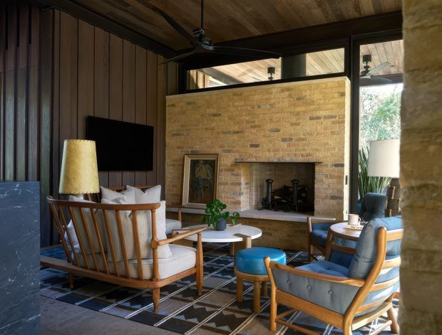 A warm, dark-toned living room with a prominent exposed brick fireplace, timber-lined ceiling and vertical timber wall panelling. Mid-century inspired timber-framed lounge chairs and a sofa surround a round coffee table, with a large geometric rug, television, lamps and indoor plants. Clerestory glazing and a tall glazed opening bring in natural light, while a ceiling fan hangs above the seating area.