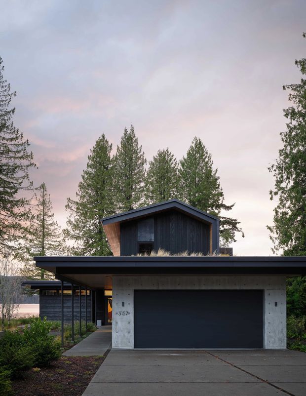 Exterior view of a contemporary two-storey house with a broad low-slung flat roof and integrated carport. The ground level features a raw concrete garage facade and dark garage door, while the upper gabled volume is clad in dark vertical timber. A paved driveway, sheltered walkway, landscaped planting, and mature trees surround the home.