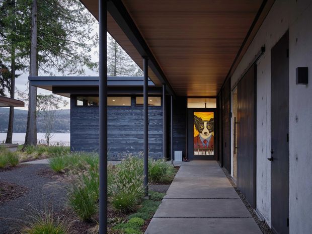 Covered exterior walkway leading through a landscaped courtyard toward a modern residence. The composition features slim dark steel posts, a warm timber-lined soffit, concrete paving, dark horizontal wall cladding, and a distinctive colourful glazed entry door. Trees and a body of water or distant landscape are visible beyond the building.