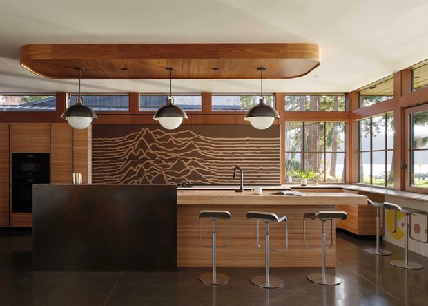 A warm, spacious mid-century-inspired kitchen with extensive timber cabinetry and framing, a large central island, dark stone surfaces, three pendant lights, and bar seating. Floor-to-ceiling windows and clerestory glazing bring in abundant natural light and views of trees and water beyond. A sculptural timber feature panel with a mountain-like linear relief forms the backsplash focal point.