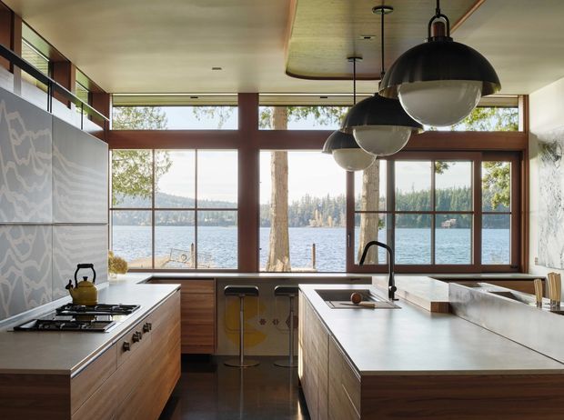 A warm, mid-century-inspired kitchen with extensive timber cabinetry, a large central island, and a gas cooktop along the left wall. Floor-to-ceiling timber-framed windows overlook a lake and wooded shoreline. Three oversized black dome pendant lights hang above the island, while a patterned feature splashback adds visual interest.