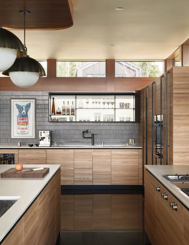 A contemporary galley-style kitchen with extensive warm timber cabinetry, dark grey vertically ridged wall tiles, pale stone benchtops, and a built-in open display cabinet stocked with glassware and bottles. Two kitchen islands frame the view, with a cooktop and integrated appliances on the right and a sink, espresso machine, and preparation area along the rear wall. Large horizontal clerestory windows bring in natural light, while dark sculptural pendant lights hang beneath a timber ceiling feature. A framed illustrated print adds a small decorative accent.