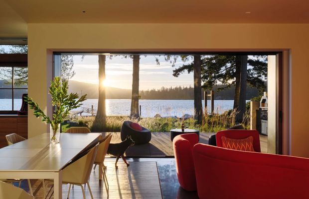 Warm, contemporary open-plan interior with a dining area and living room arranged around a large picture window overlooking a lake or waterfront landscape. The room features a pale dining table with light timber chairs, sculptural dark lounge seating, and a bold red sofa and armchair. Timber flooring, warm neutral walls and ceiling, and extensive glazing create a bright connection to the outdoors.