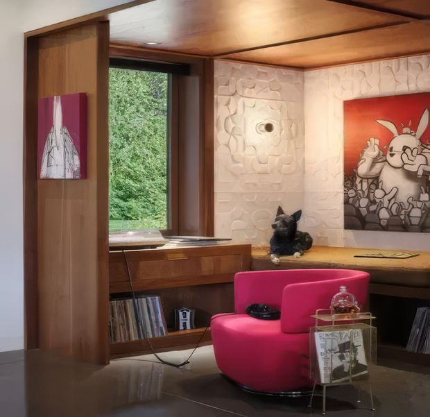 A compact, richly detailed mid-century-inspired study or living space with extensive warm timber wall and ceiling surfaces, built-in desk and shelving, a large window overlooking greenery, sculptural textured wall panels, and a vivid pink upholstered lounge chair. Artwork, records, and small decorative objects add an eclectic character.