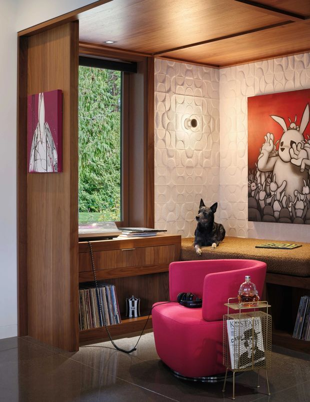 A richly detailed mid-century-inspired living room or media nook featuring warm timber wall and ceiling panelling, built-in shelving and a window seat, textured geometric wall tiles, a vivid pink lounge chair, and colourful artwork. Vinyl records and a small drinks trolley reinforce the retro lounge character. A dog is seated on the built-in bench.