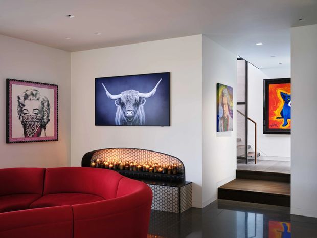 A stylish contemporary living room with a large curved red sectional sofa, polished dark flooring, and a dramatic built-in fireplace lined with patterned tile and candles. The walls display bold artwork, including a Highland cow portrait, while steps lead to a raised hallway or adjoining level with additional artwork and a modern stair railing. Recessed ceiling lighting and crisp white walls create a refined gallery-like interior.
