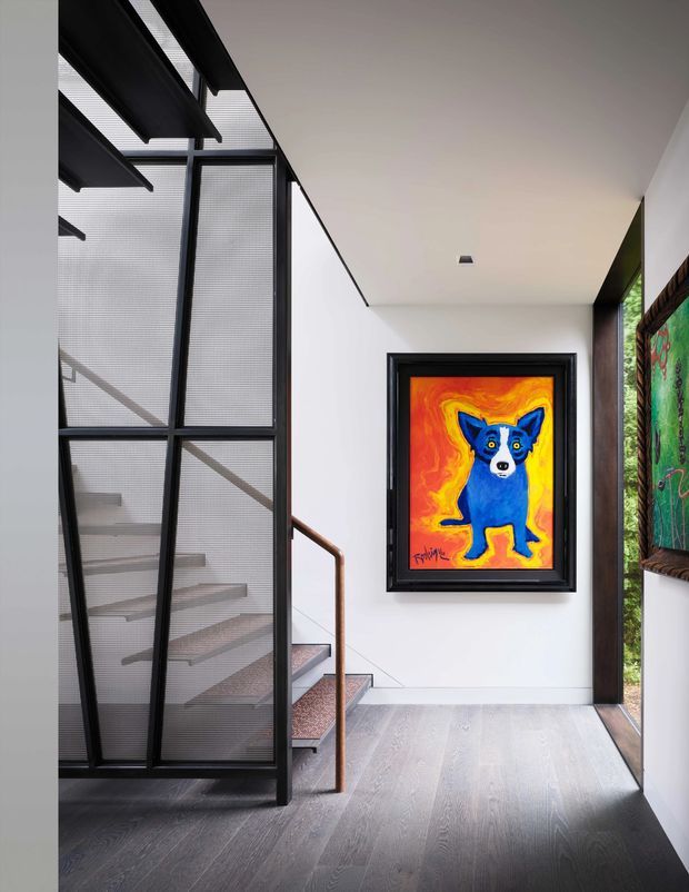 A modern interior landing or hallway with a dark timber floor, white walls and ceiling, black-framed textured glazing enclosing a staircase, and a slim bronze-toned handrail. A brightly coloured framed artwork is mounted on the wall, with additional artwork visible beside a narrow glazed opening.