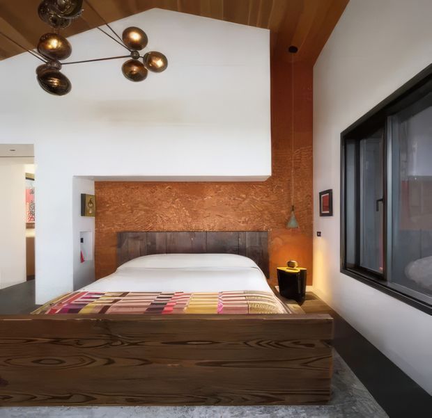 A compact, boldly designed bedroom featuring a low platform bed with a substantial dark timber frame and headboard. The bed is set against a textured warm terracotta feature wall, with white walls and a sculptural white overhead volume. A large black-framed window occupies the right wall, while a modern cluster pendant light, small bedside table, and colourful folded textile add visual interest. The room has a warm, eclectic contemporary character.