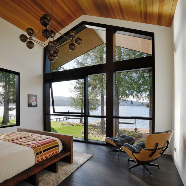A mid-century-inspired bedroom with a timber-lined sloped ceiling, dark stained timber flooring, and expansive black-framed glazing overlooking a lakeside setting. The room contains a low timber bed with a colourful geometric textile, a sculptural multi-globe pendant light, and a classic lounge chair with ottoman. White walls and large windows create a bright connection to the outdoors.
